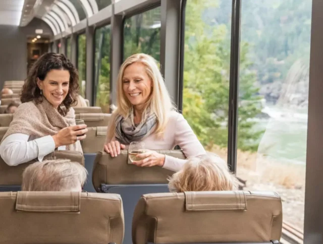 Two women stand smiling with drinks inside a train, engaging in conversation with two seated individuals. The train windows reveal a view of greenery outside.