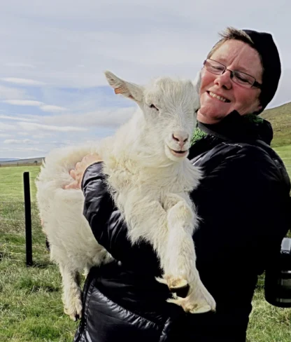 Two people in outdoor clothing, one holds a white goat and smiles. They're standing in a grassy area with a fence and hills in the background.