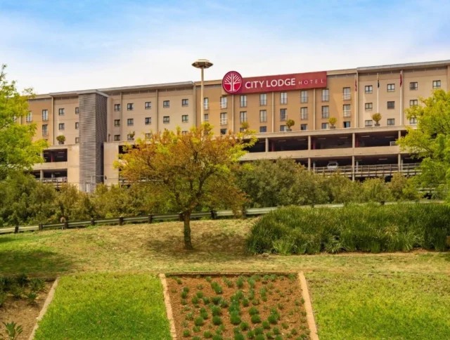 A multi-story City Lodge hotel with a red logo sign, surrounded by green trees and neatly landscaped gardens in the foreground.