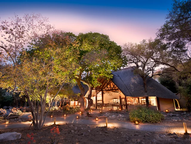 A cozy lodge with a thatched roof surrounded by trees is illuminated at dusk. The path leading to the entrance is lined with small lights.