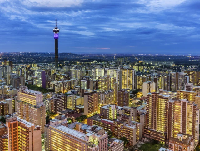 Aerial view of a cityscape with numerous buildings and a prominent tower. The sky is cloudy, and the city is illuminated, suggesting evening or night time.