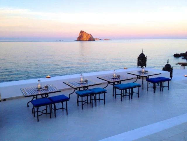 Outdoor dining area with tables and blue chairs overlooking a calm sea at sunset, with a small rocky island in the distance.