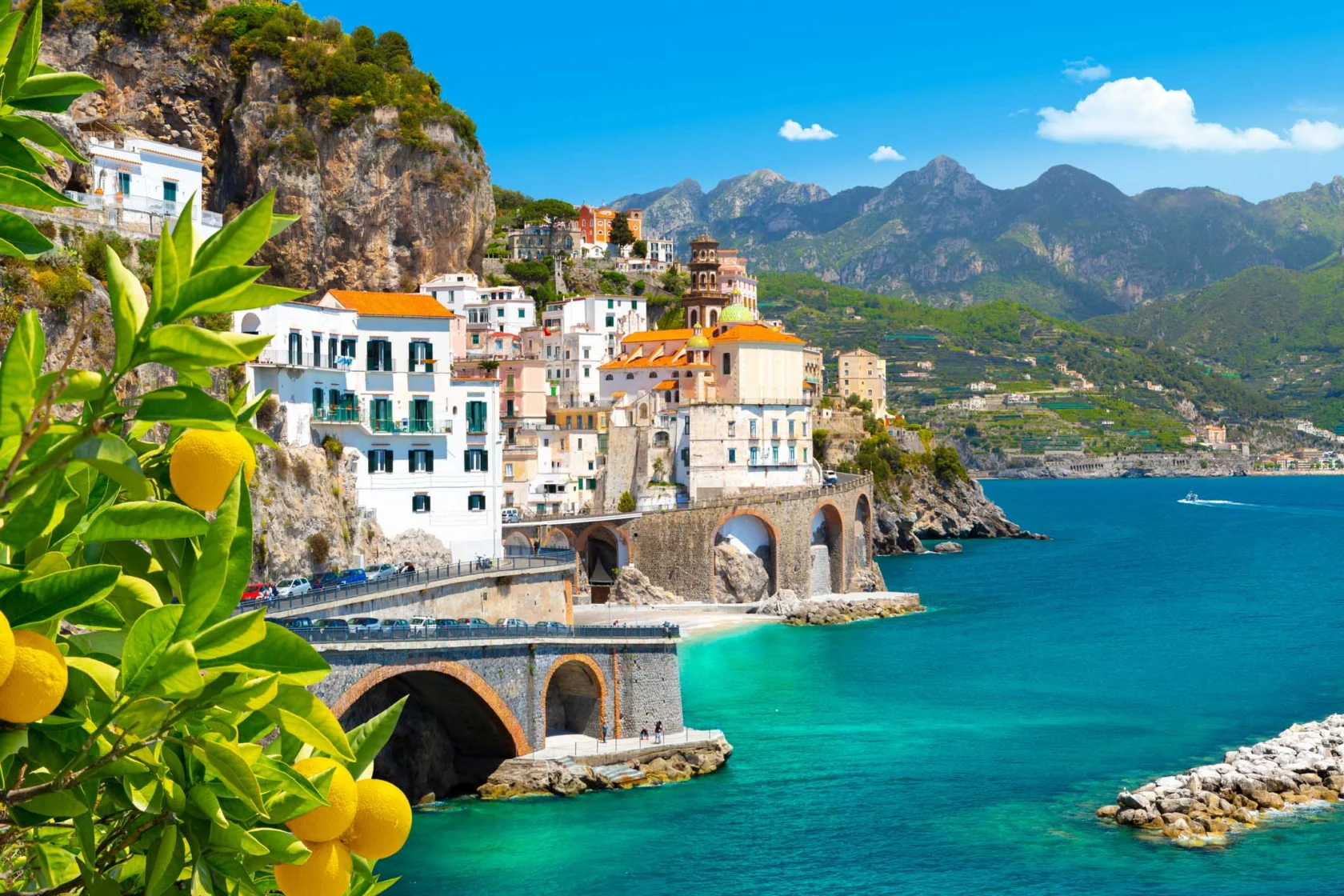 Coastal town with colorful buildings on a cliff, surrounded by clear blue sea and mountains. Lemons hang from a tree in the foreground.