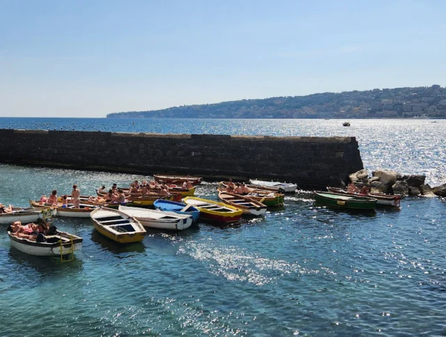 Boats are moored in a small harbor next to a stone wall with people swimming nearby. Background shows a sunny coastline with buildings on a distant hill.