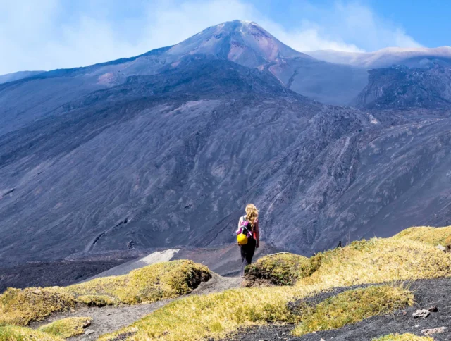 A person with a backpack walks on a path surrounded by sparse vegetation, with a large, rocky mountain in the background.