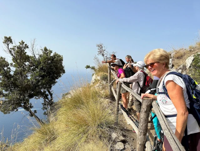 A group of people stand on a wooden railing platform overlooking a coastal cliff, with grassy vegetation and a distant view of the sea.