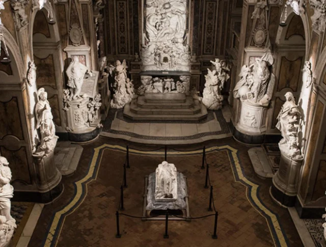 A grand hall with elaborate marble statues and sculptures lining the walls and a central sculpted figure on the floor, surrounded by a decorative, patterned floor.