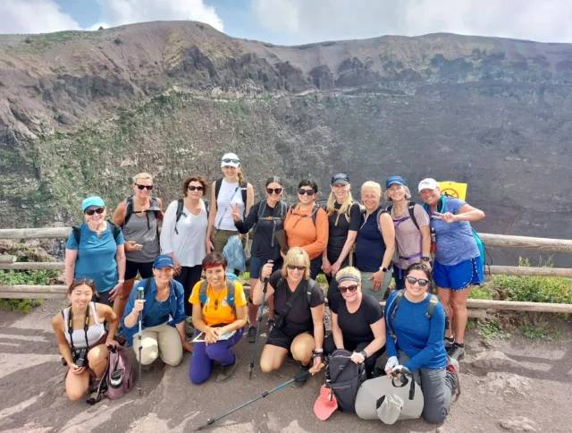 A group of people in hiking attire pose for a photo in front of a mountainous crater, with a wooden fence in the background.