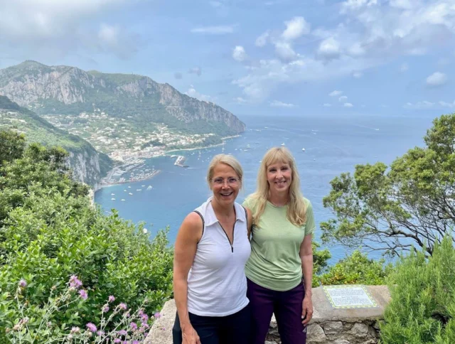 Two people smiling with a scenic view of a coastal landscape, featuring cliffs, greenery, and the ocean under a partly cloudy sky.
