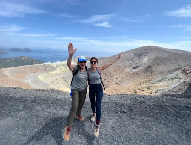 Two people in hiking gear pose with arms raised on a rocky mountain terrain under a clear blue sky.