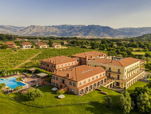 Aerial view of a large villa with a red-tiled roof surrounded by greenery and mountains, featuring a swimming pool and shaded areas.