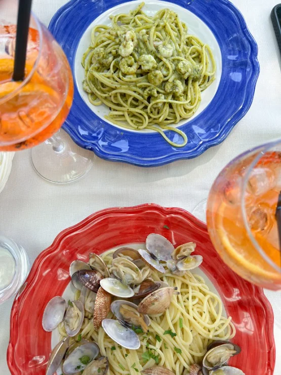 Two plates of pasta: one with clams on a red plate and one with pesto and shrimp on a blue plate. Two glasses of orange spritz are visible.