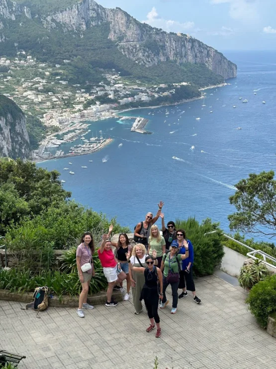 A group of people pose on a terrace overlooking a coastal town and cliffs, with a view of the sea and boats in the background.