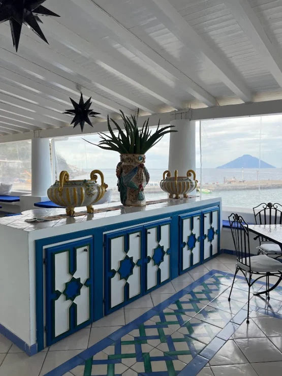 A Mediterranean-style terrace with blue and white decor, ceramic pots, and a mountain view through large windows.