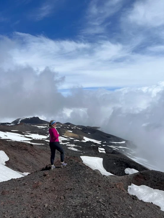 Person standing on a rocky, snow-dotted mountain slope with a cloudy sky in the background.