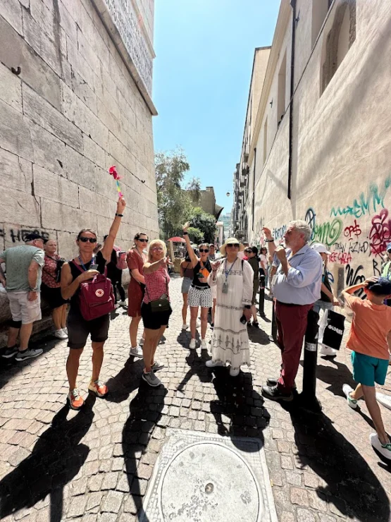 A group of tourists stands in a narrow, graffiti-covered alley, some holding red flowers, on a sunny day.