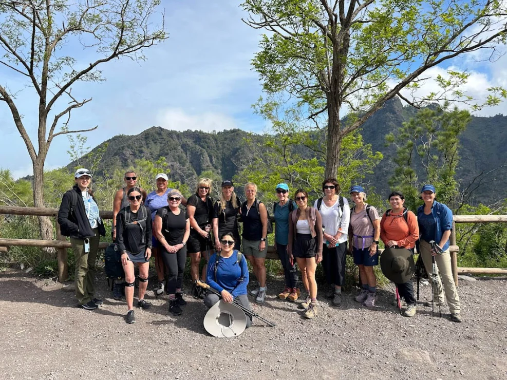 A group of people standing in front of a mountain landscape with trees and a wooden fence.