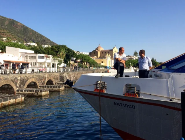 Two people standing on a boat docked near a stone pier, with buildings and a hill in the background on a sunny day.