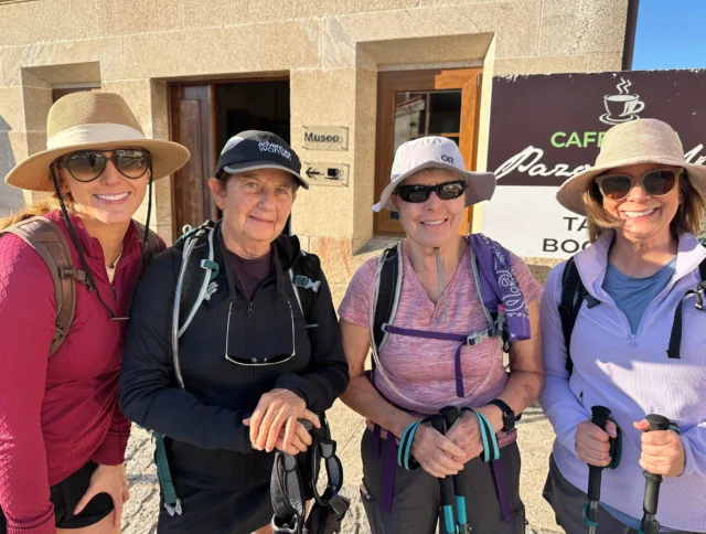Four women with sun hats and hiking gear stand outside a building with a "Café" sign.