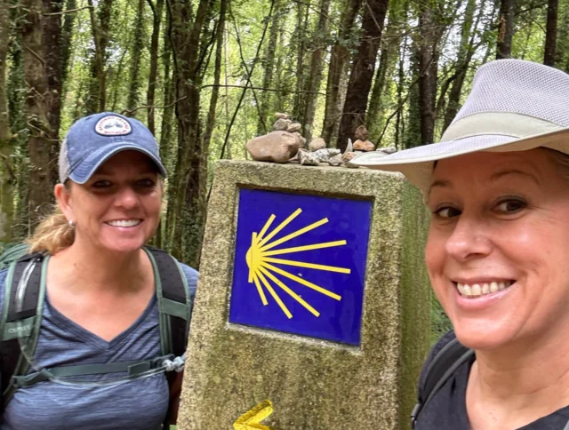 Two people smiling beside a Camino de Santiago marker in a forest, with a yellow arrow and shell symbol displaying "Km 110" on a stone post.