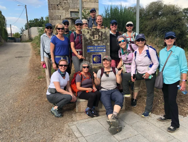 A group of 14 people posing in front of a Camino de Santiago signpost, wearing casual hiking attire and smiling.
