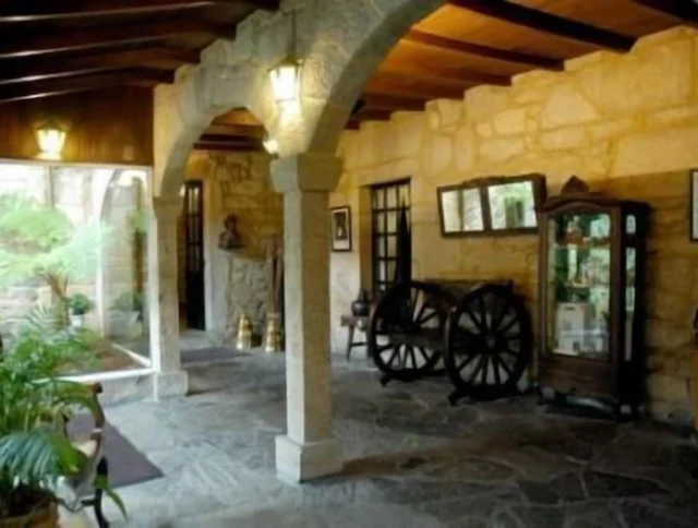 Stone interior with wooden ceiling, arched columns, and large windows. Decor includes plants, a wagon wheel, statues, and a cabinet displaying items.