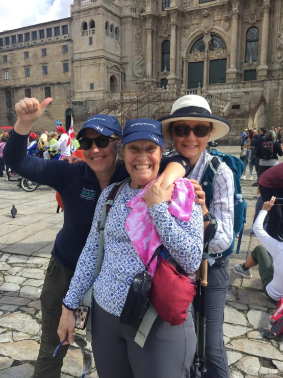 Three people smiling and posing in front of an old stone building. Two wear hats and sunglasses, and one holds a pink garment. There are people and pigeons in the background.