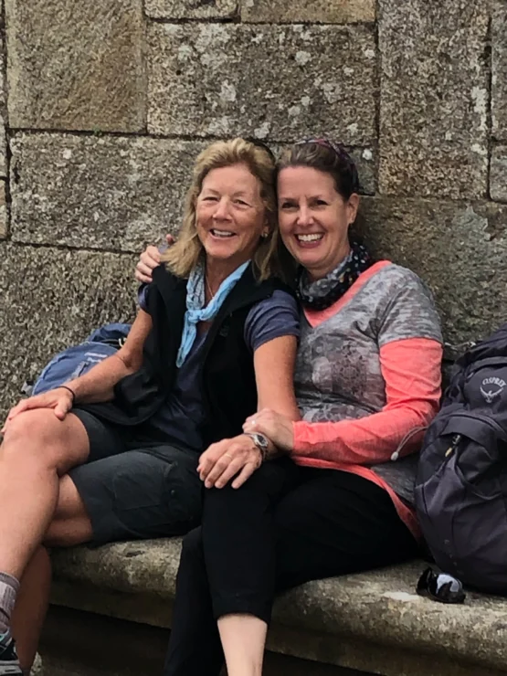 Two women sitting on a stone bench, smiling and embracing. They both have backpacks next to them.
