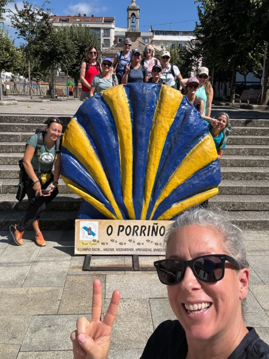 A group of people posing in front of a large blue and yellow shell sculpture with a sign reading "O Porriño" on steps of an outdoor plaza. One person in front is making a peace sign.