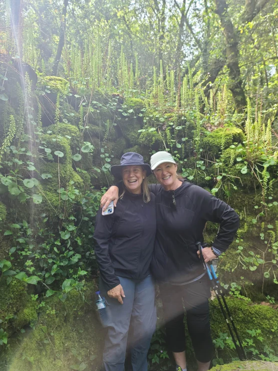Two people wearing hats and outdoor clothing pose together on a lush green hiking trail, surrounded by ferns and mossy rocks.