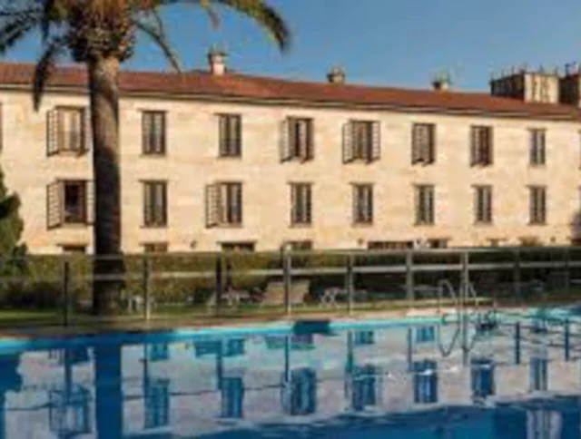 Large stone building with numerous windows, surrounded by trees, reflected in a swimming pool in the foreground.