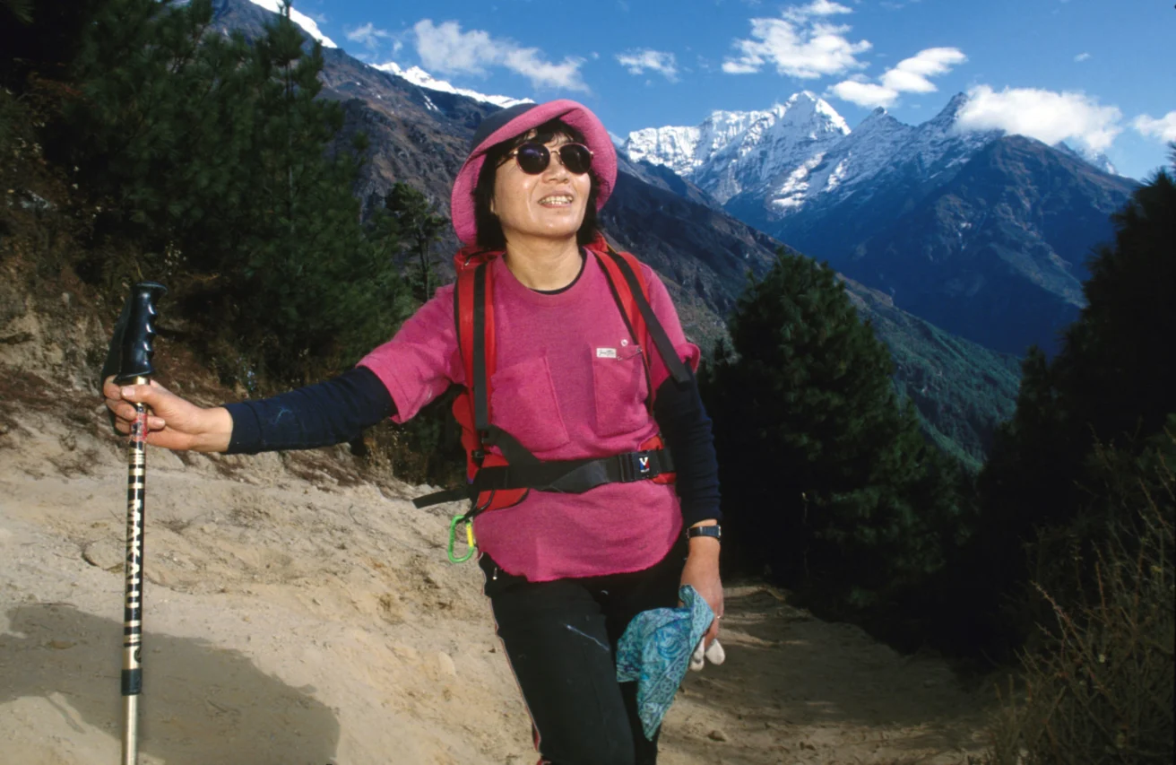 A person hiking on a mountain trail, wearing a pink hat and sunglasses, holding a walking stick. Snow-capped mountains and trees are in the background.