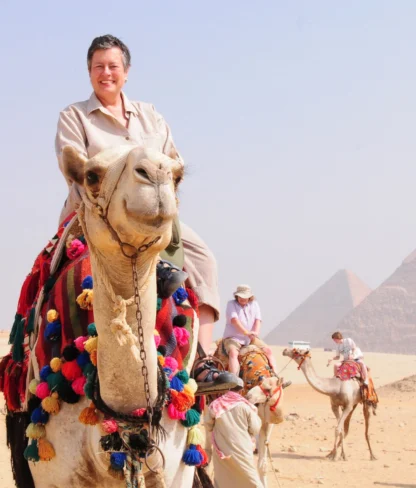 A person rides a decorated camel in a desert near pyramids, with others on camels in the background.
