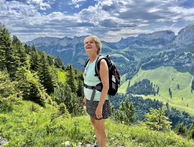 A person stands on a grassy mountain trail, wearing a backpack and looking up at the sky. The background features green hills and a cloudy sky.