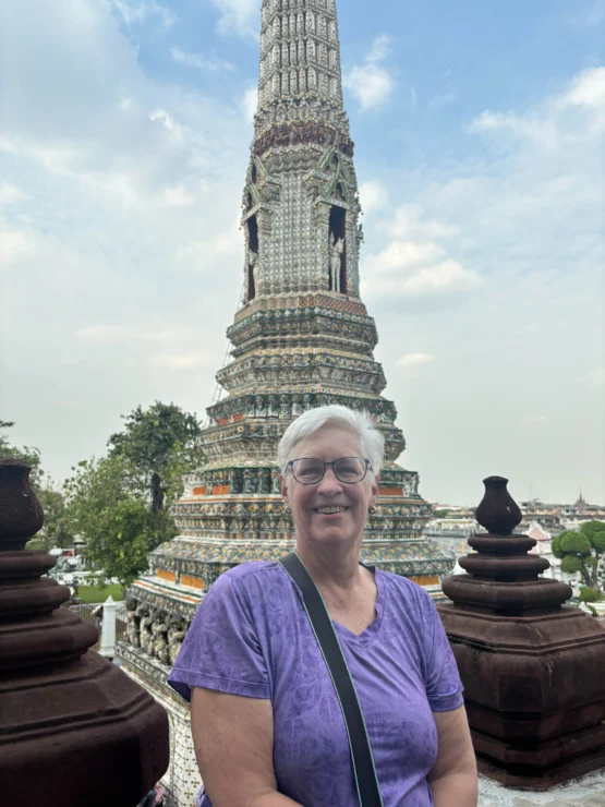 A person in a purple shirt stands smiling in front of a tall, ornate tower with intricate designs. The sky is partly cloudy.