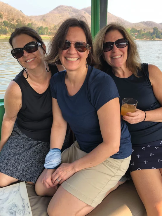 Three people wearing sunglasses sitting together on a boat, with a lake and hills in the background. One person holds a drink.
