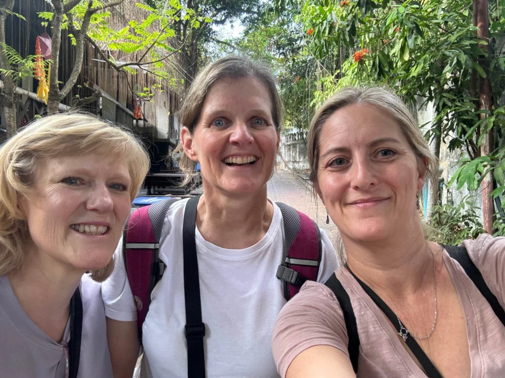 Three people smiling, posing for a selfie outdoors with trees and a fence in the background.