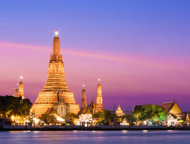Wat Arun temple with illuminated spires at dusk, set against a vibrant purple and pink sky, viewed from across a river.