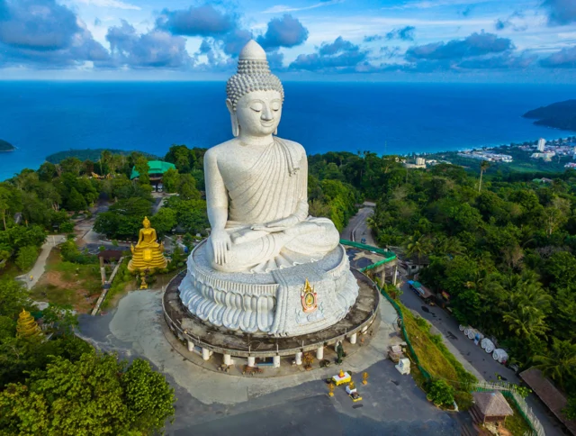 Aerial view of the Big Buddha statue on a hill surrounded by lush greenery, with a coastal town and ocean in the distance under a partly cloudy sky.