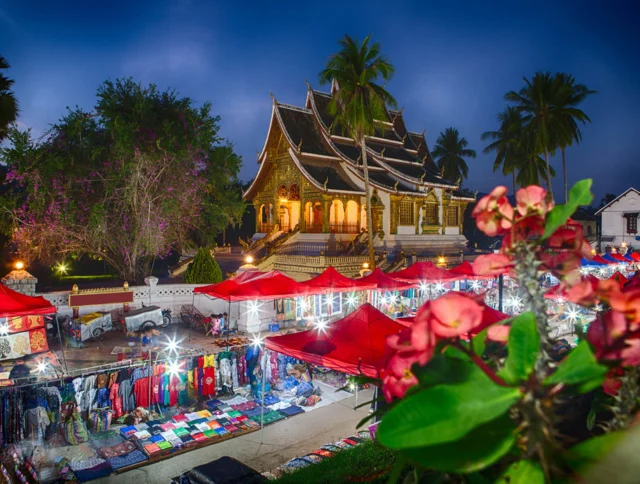 Night market with colorful stalls and red canopies in front of an illuminated traditional temple. Palm trees and flowering plants are visible against a dusky blue sky.