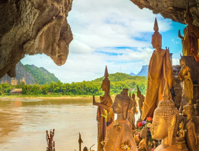 A collection of Buddha statues overlooks a river through a cave opening, surrounded by offerings and vibrant greenery.