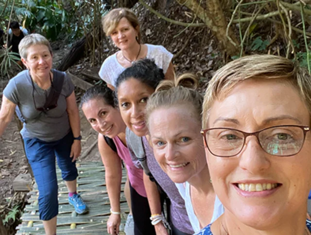Group of six women smiling while standing on a bamboo bridge in a forested area.
