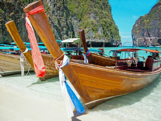 Long-tail boats anchored on a sandy beach with clear turquoise water, surrounded by towering limestone cliffs under a bright blue sky.
