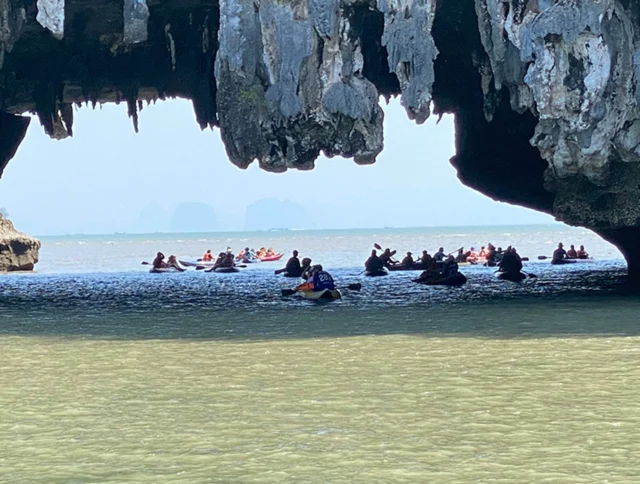 People in kayaks paddle under a rocky overhang in a calm body of water.