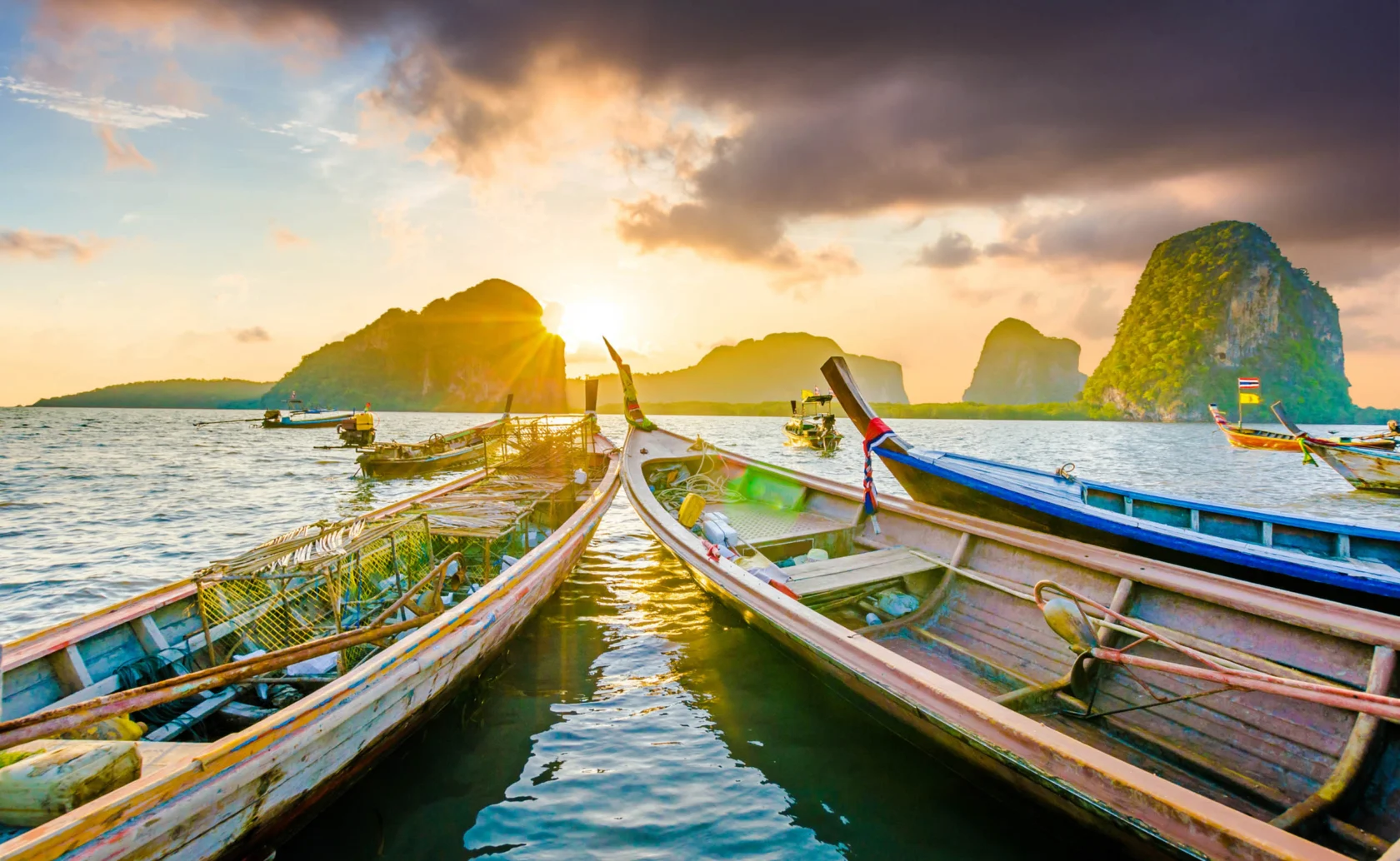 Several long-tail boats on water with dramatic sky and rocky islands in the background at sunset.