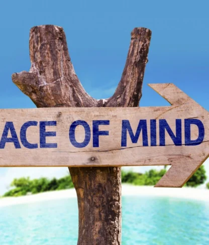 Wooden sign with "Peace of Mind" written on it, mounted on a tree branch, pointing towards a beach with clear blue sky.