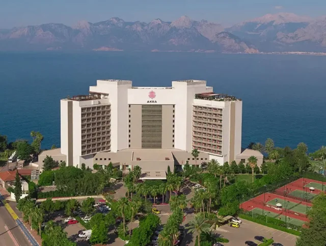 A large hotel building by the seaside, surrounded by greenery and sports facilities, with mountains in the background.