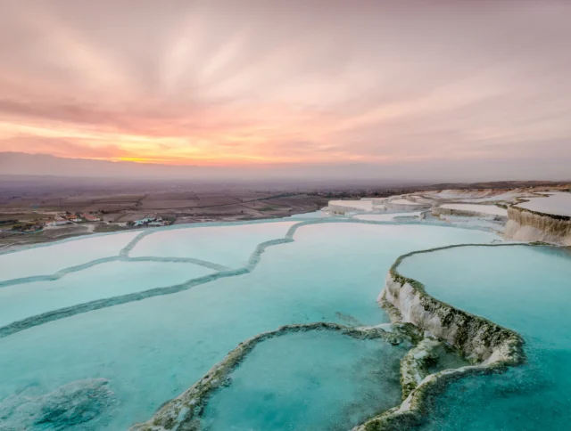 Terraced pools with turquoise water and white sediment formations at sunset, with a vast landscape in the background.