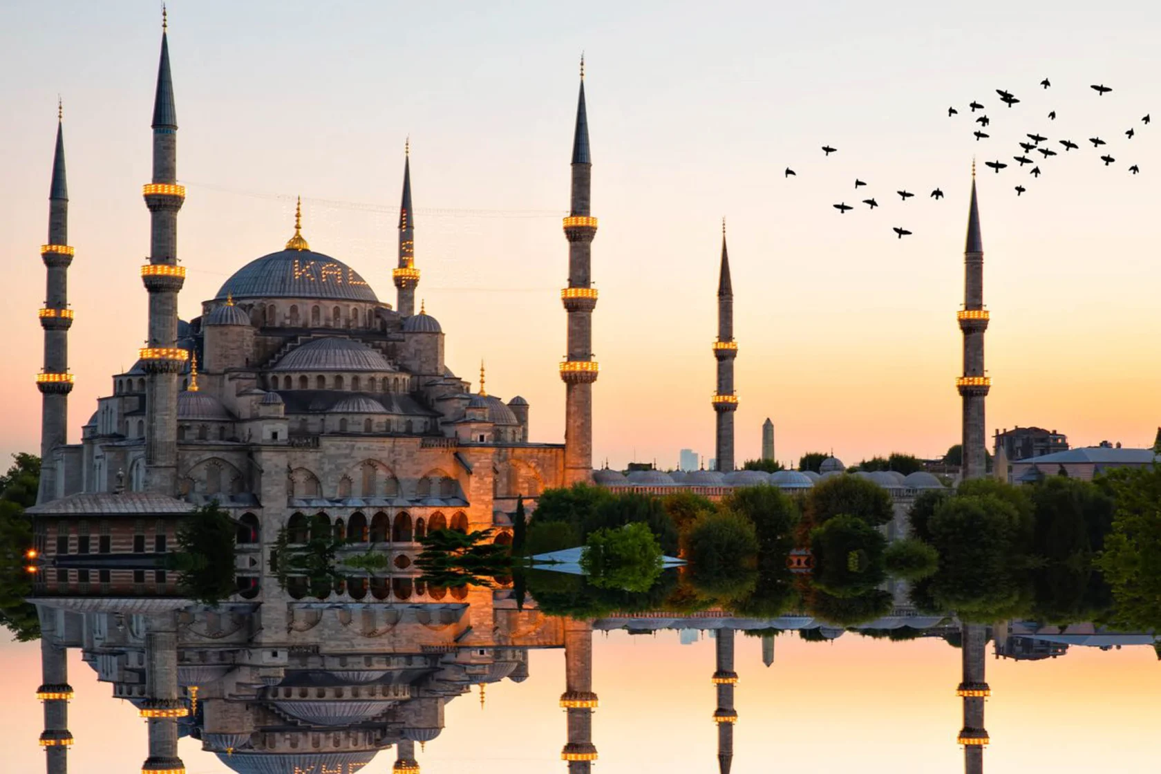 The Blue Mosque at sunset with birds flying in the sky, reflected in tranquil water, surrounded by greenery.