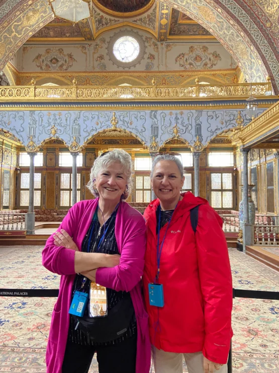 Two people smiling and posing inside an ornate, historic room with decorative patterns, high ceilings, and large windows.